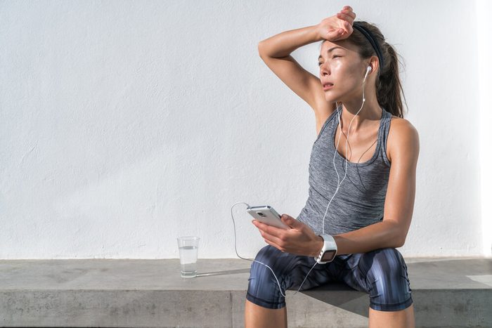 Tired fitness woman sweating taking a break listening to music on phone after difficult training. Exhausted Asian runner dehydrated feeling exhaustion and dehydration from working out at gym.
