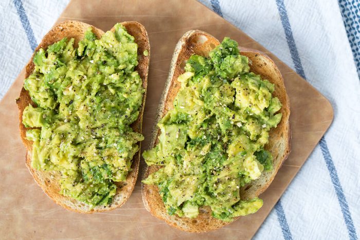 Heart healthy diet; Over head avocado toast on slices of toasted French loaf bread topped with ground lemon pepper and sprinkles of kosher salt on wood cutting board on top blue and white striped linen cloth