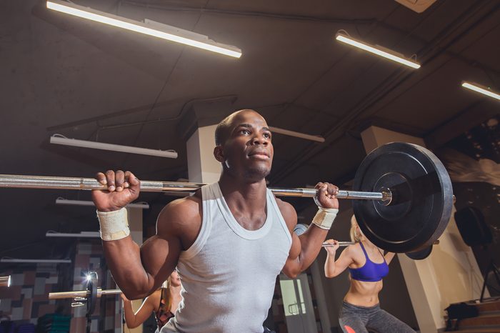 Fit young people lifting barbells over their heads looking focused, working out in a gym with other people
