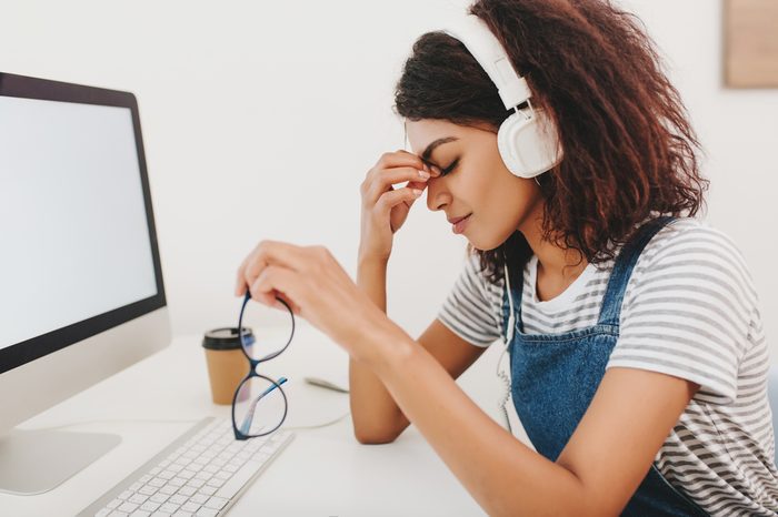 Woman in headphones with headache sitting beside computer and holding glasses in hand.