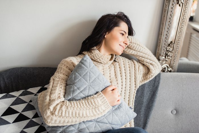 Horizontal shot of beautiful young Caucasian woman sitting on grey sofa near the Christmas tree, holding a pillow and looking at one side with a lovely smile. Christmas and New Year mood.