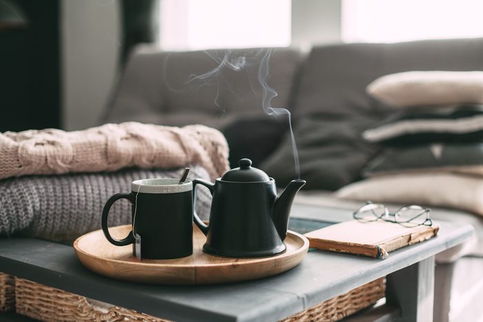 Still life details in home interior of living room. Sweaters and cup of tea with steam on a serving tray on a coffee table. Breakfast over sofa in morning sunlight. Cozy autumn or winter concept.