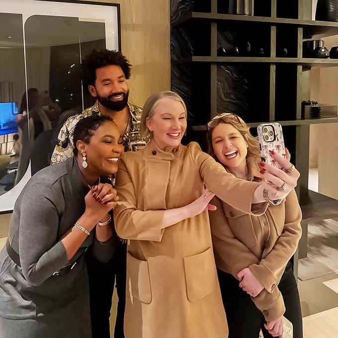 Four people are gathered in a well-lit room, posing for a selfie. They are smiling and standing close together. A woman in a beige coat is holding the phone. The background shows a modern interior with shelves and a reflection in a mirror.