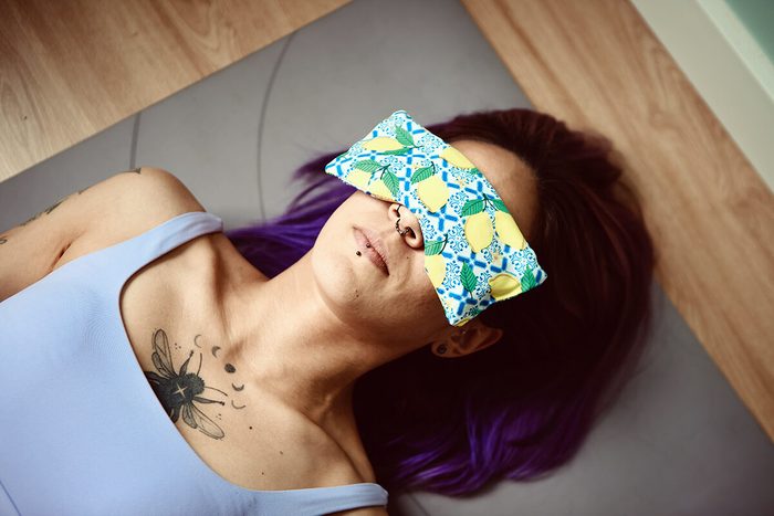 Person lying on a yoga mat with a colorful eye pillow covering their eyes. They have long, purple hair and tattoos on their shoulder and chest. The background features wooden flooring.