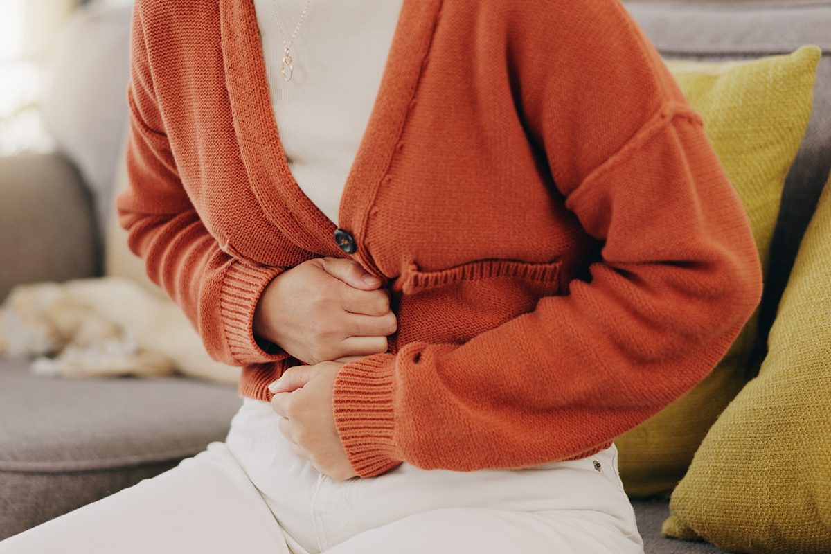 Stomach Ache, Cramps And Hands Of Woman With Abdomen Pain Due To Constipation, Menstruation Or Ibs Issue. Sick, Home And Person Suffering And Holding Belly In A House Lounge, Couch And Living Room